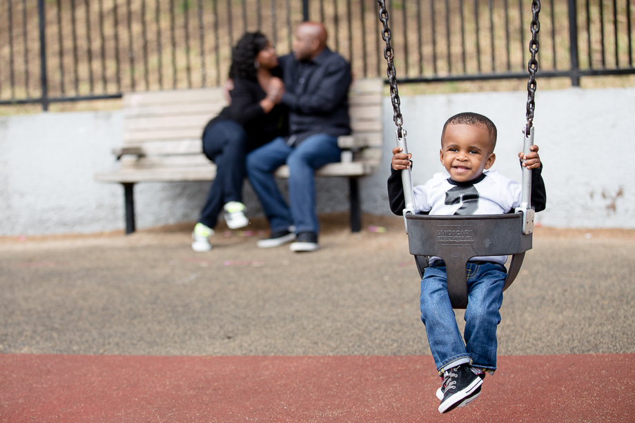 Two-year-old Journey on a swing set with his loving parents on a park bench