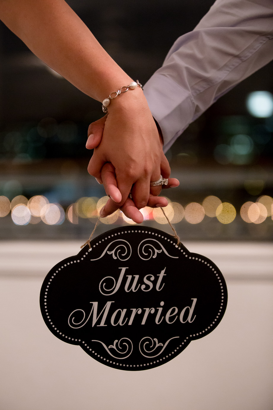 Newlywed couple holding Just Married sign and showing off ring in front of a beautiful line of blurred lights