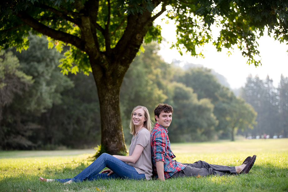Engaged couple sitting back-to-back under a tree in Palo Alto