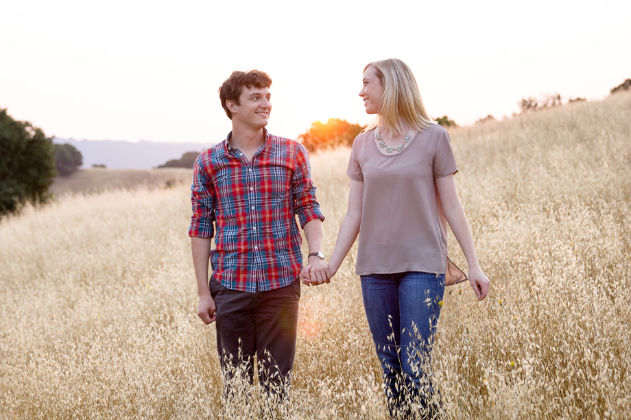 Happy couple holding hands and looking at each other as they walk in grassy field under a warm sunset