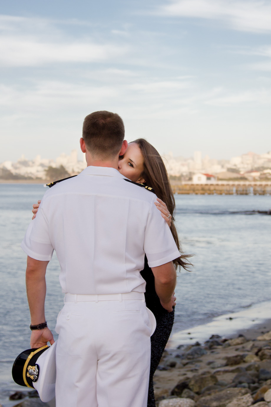 Kissing his neck peeking over his shoulder as he holds his US Navy cap and looks off into San Francisco Bay