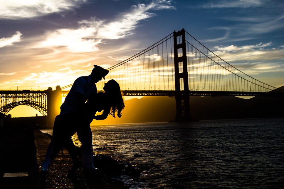 Silhouette engagement photo of US Navy officer and fiancee doing The Dip over water in front of San Francisco Golden Gate Bridge with orange, blue, pink sky