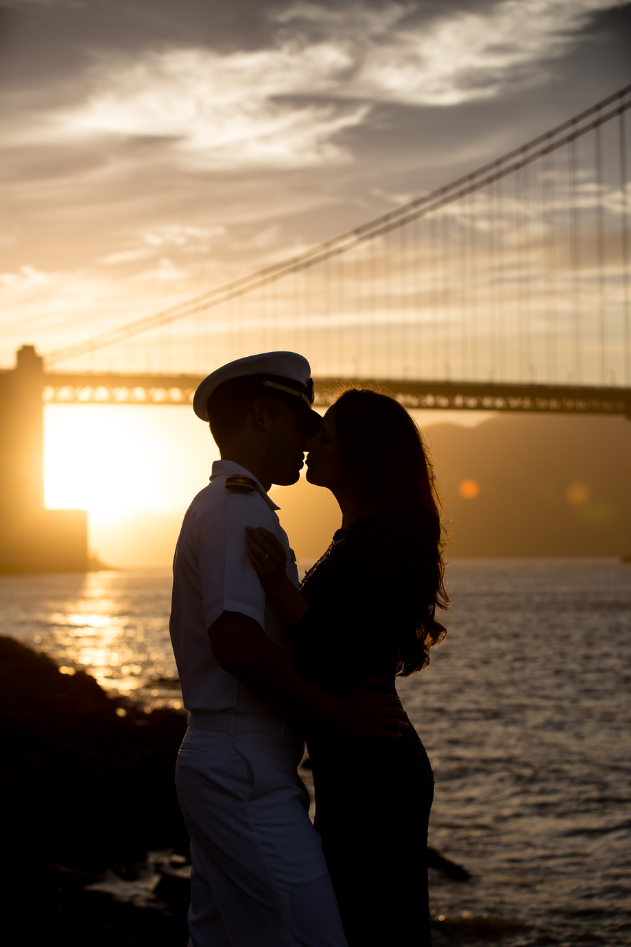 Romantic engagement photo of couple kissing at the base of San Francisco Golden Gate Bridge backlit by the setting sun showing a silhouetted heart