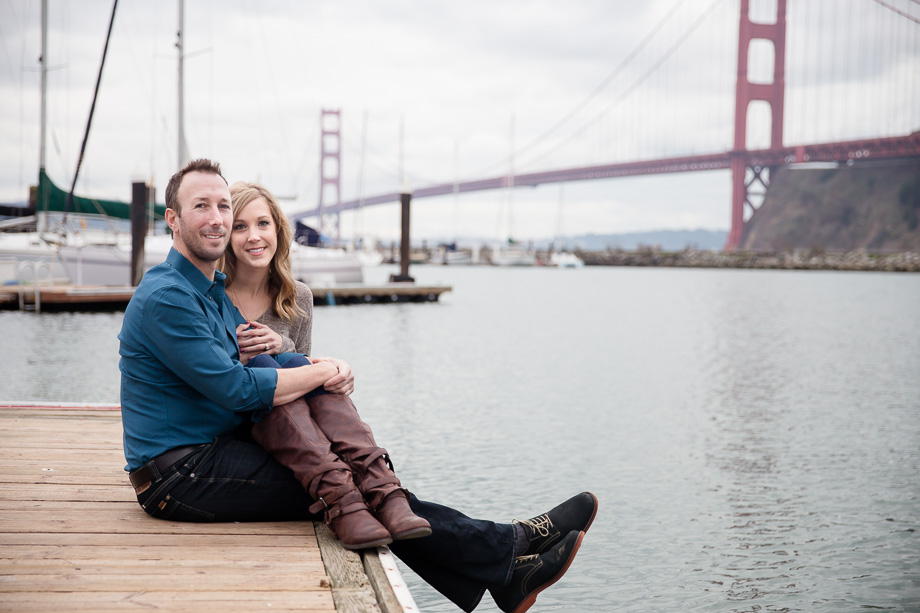 engagement photo of couple sitting at the end of a pier in front of the Golden Gate Bridge and boats