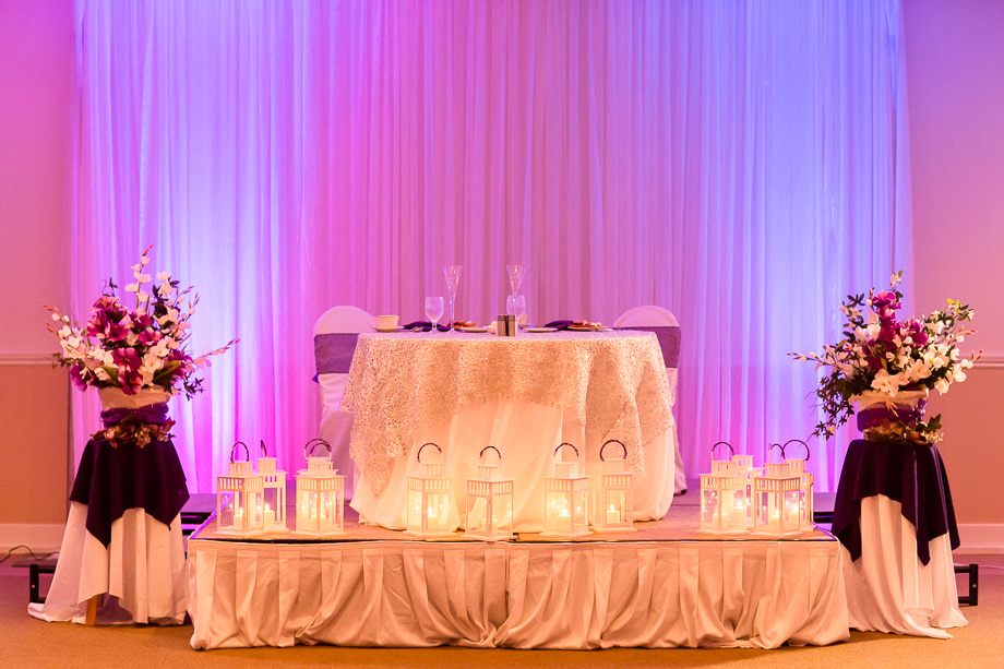 sweetheart head table at Boundary Oak Golf Club reception hall with champagne and purple floral arrangements and lighting