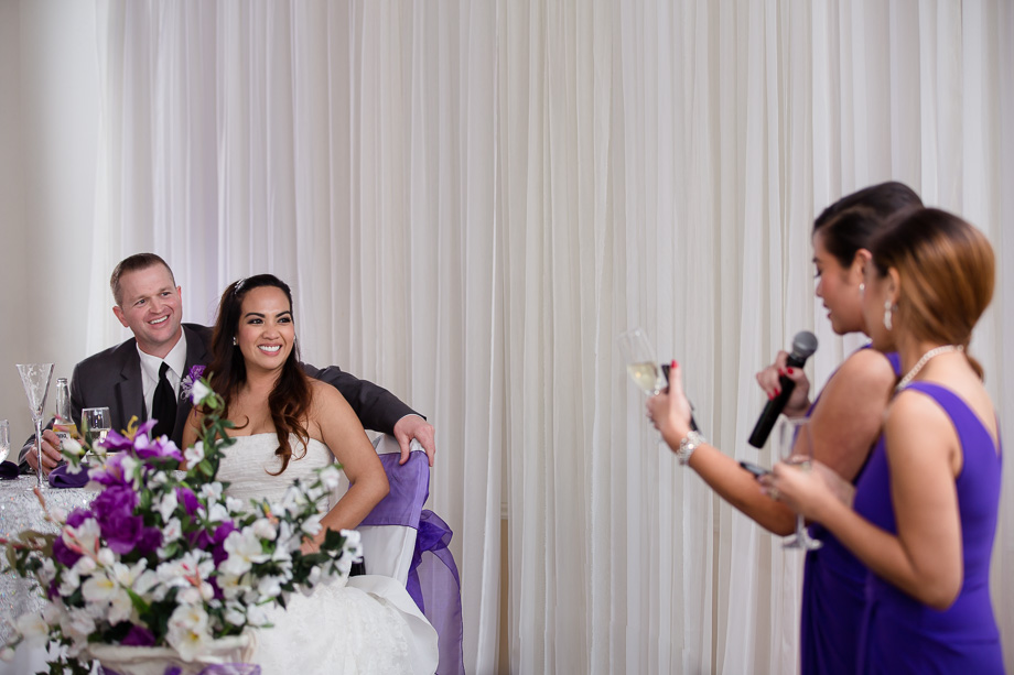 bride and groom holding champagne glasses behind floral decorations looking at the bridesmaids giving toasts