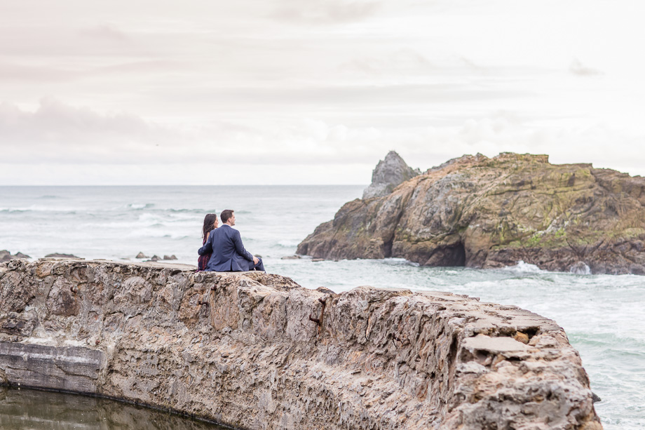 engagement photo couple sitting on Sutro Baths ruins facing the ocean
