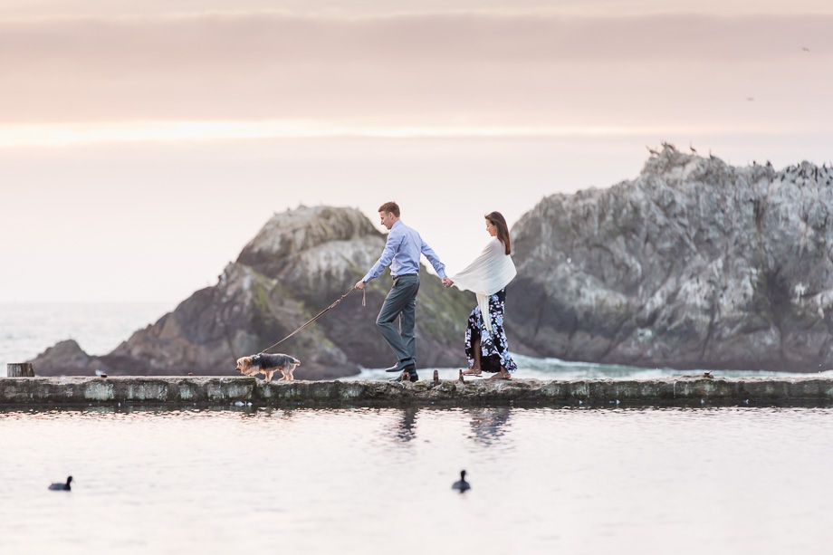 engagement photo of couple walking with their dog along the sutro baths water during sunset in San Francisco