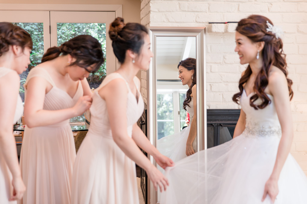 bride smiling in mirror at bridesmaid helping her with dress