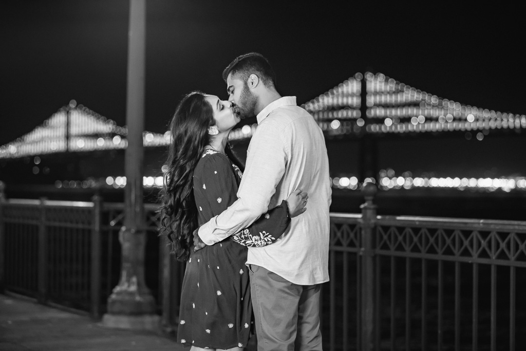 black and white engagement photo with lit Bay Bridge at night