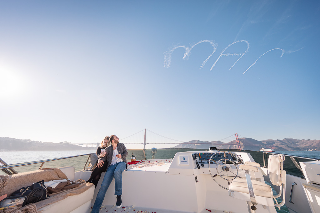 newly engaged couple sitting under marry me written in the sky in huge letters