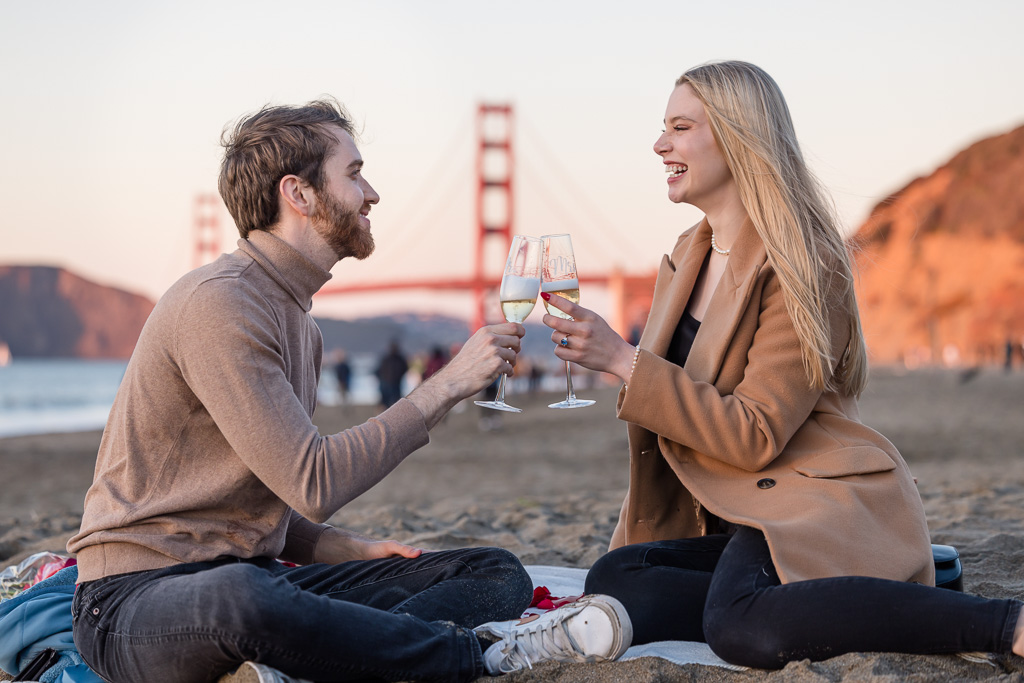 celebrating engagement with champagne glasses on the beach with the Golden Gate in the background