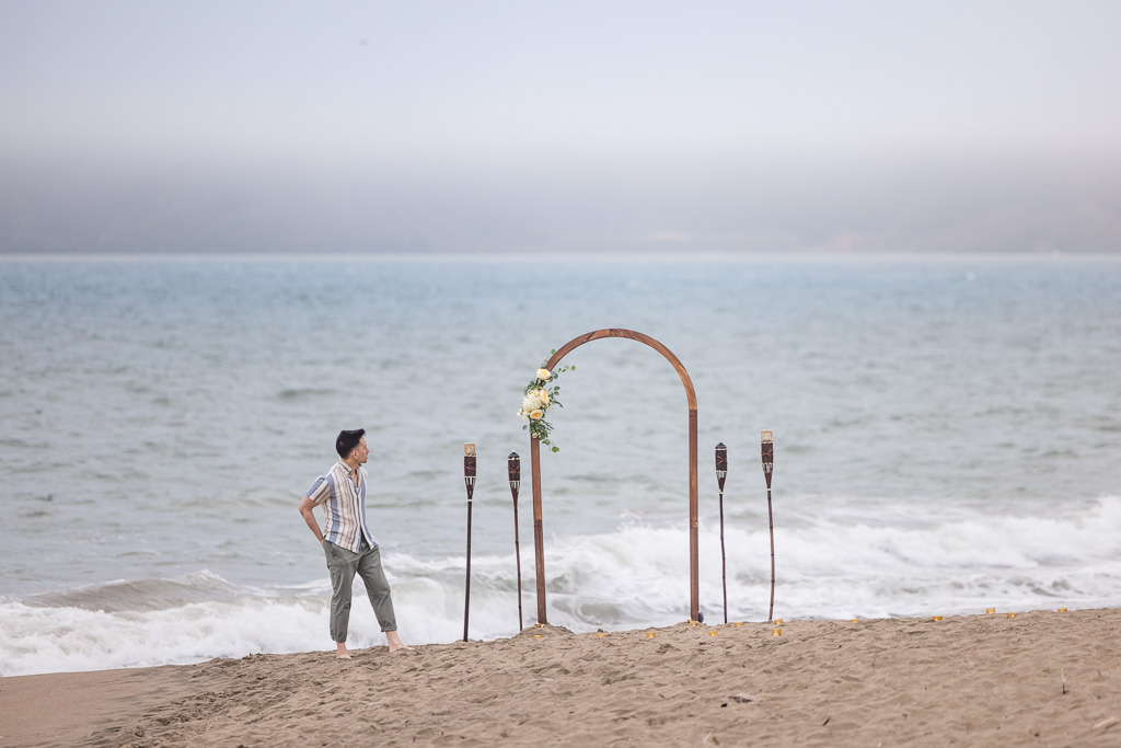 groom-to-be standing next to his DIY Baker Beach proposal arch and tiki torches