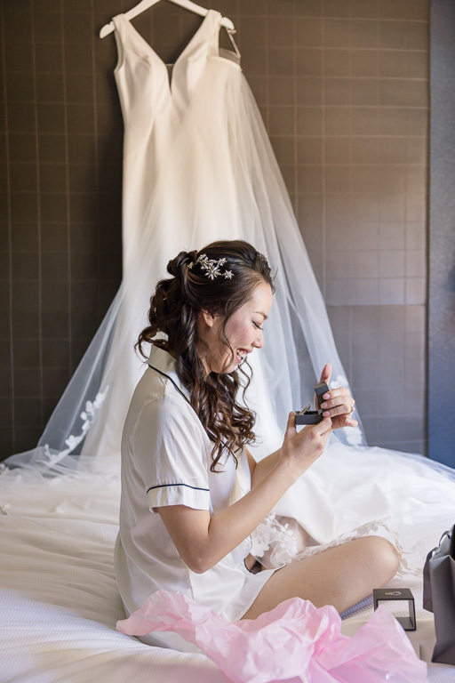 bride sitting on bed in front of her dress looking at a gift from her husband-to-be