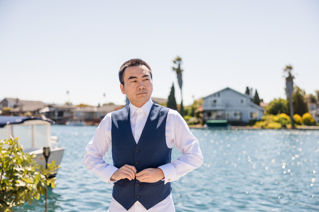 groom buttoning up vest in front of a lake