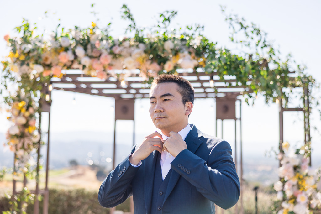 groom adjusting his tie in front of floral arch outdoors