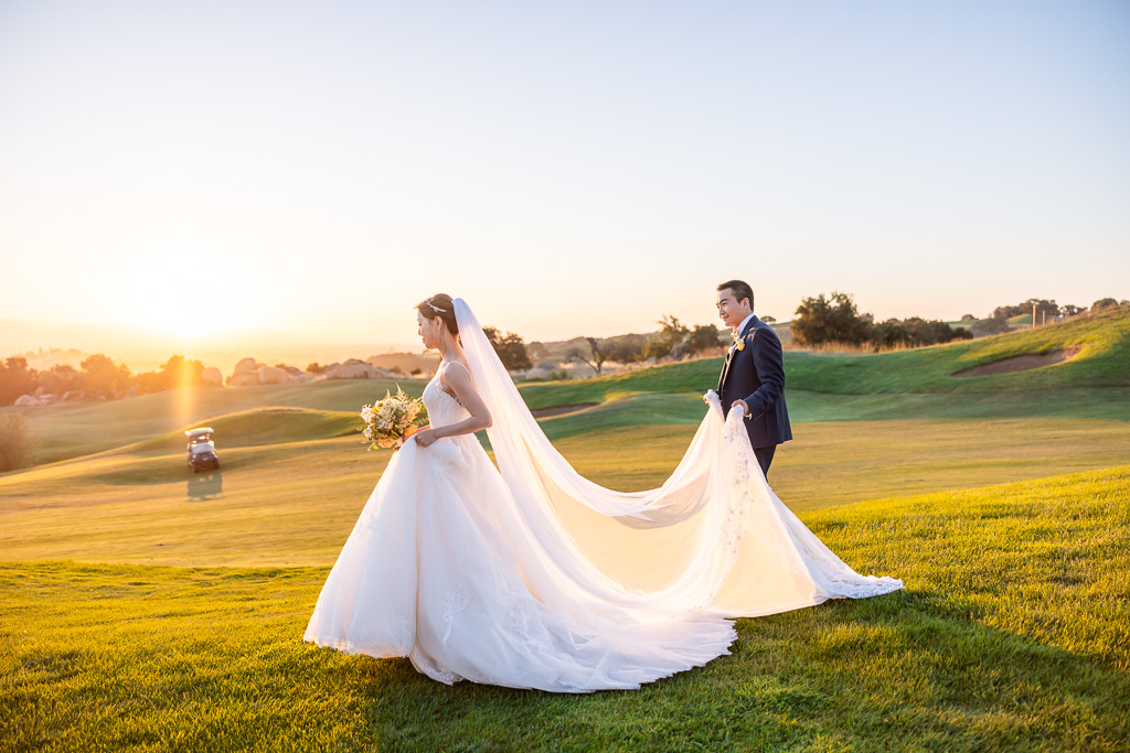 groom holding veil behind bride as they walk down a grassy hill during sunset hour