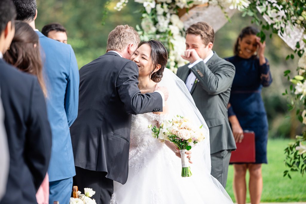 bride hugging the father of the groom with the groom having a moment in the background