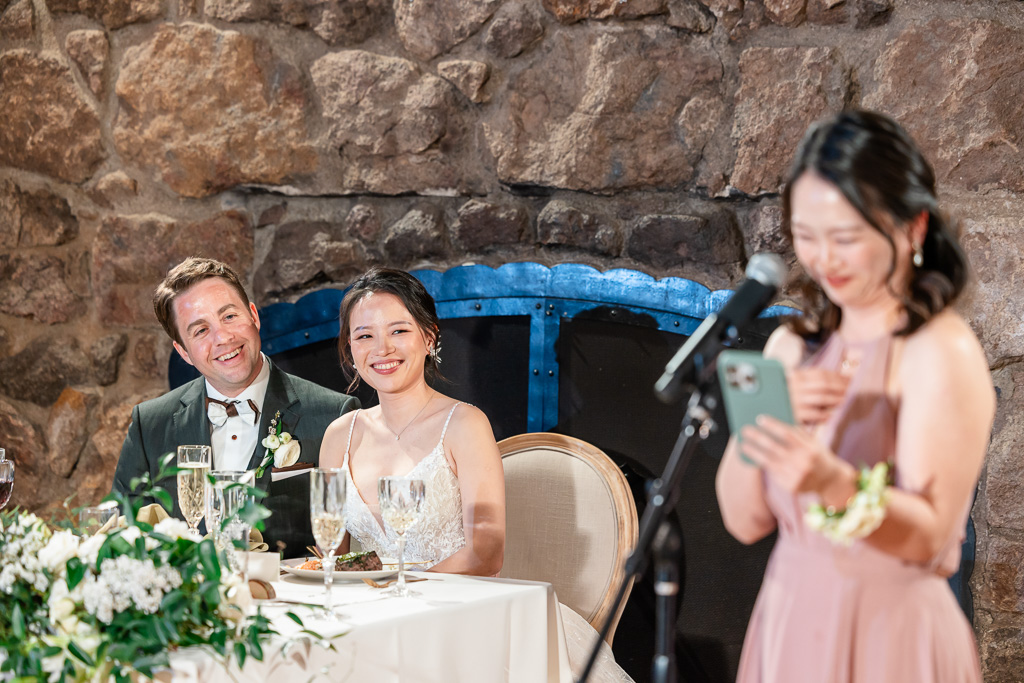 bride and groom smiling at bridesmaid giving a speech