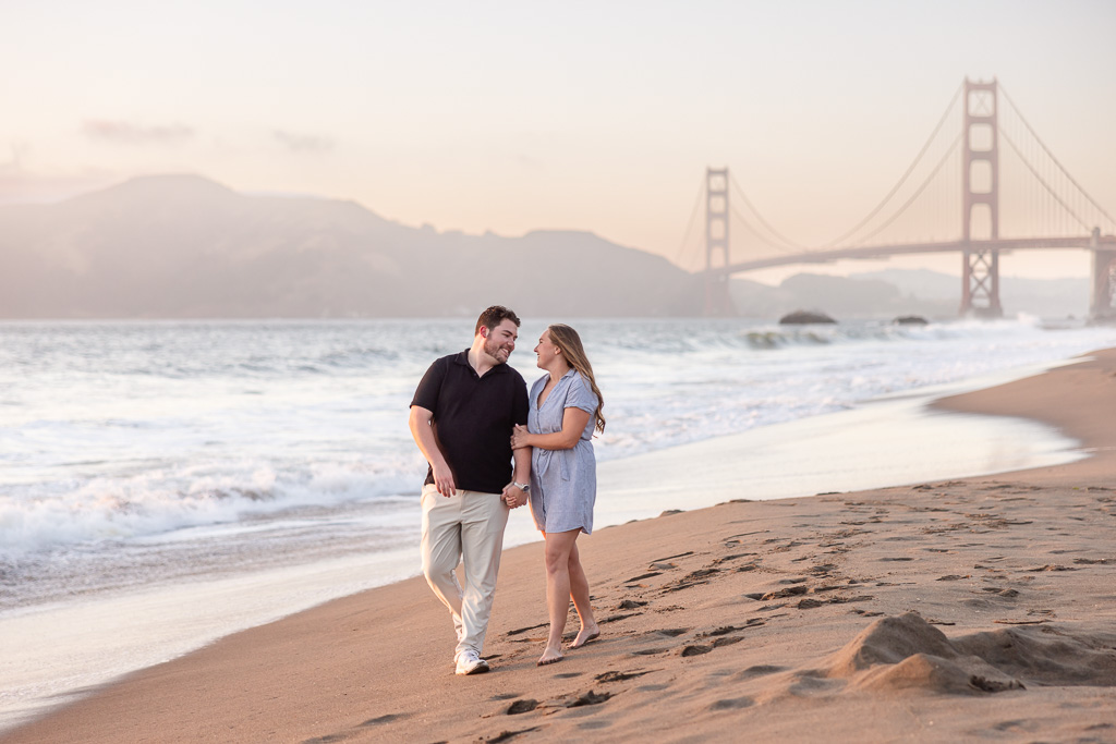 engagement photo of couple walking along the shoreline in front of Golden Gate Bridge