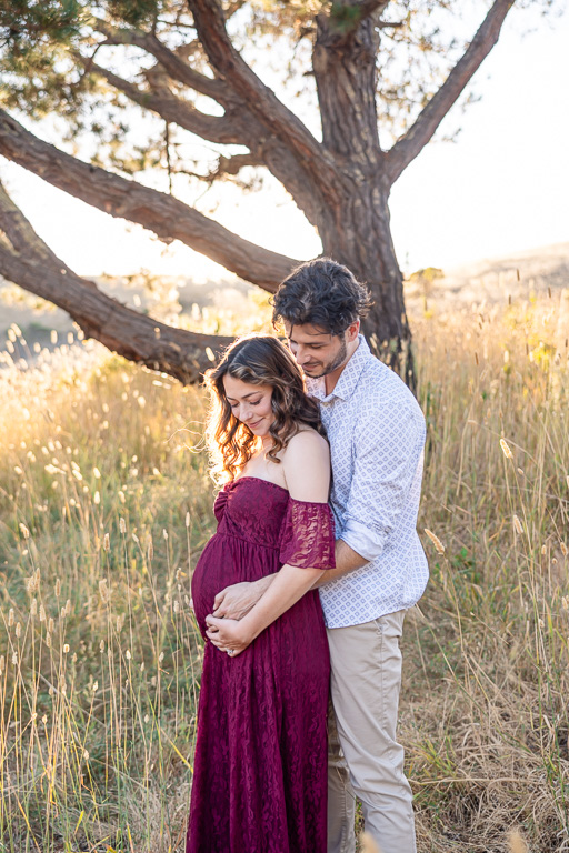 A maternity photo at sunset in the Foothills Nature Preserve, Los Altos Hills. The expecting couple stands in a golden field, with the woman in a burgundy lace maternity dress cradling her baby bump as her partner embraces her from behind.