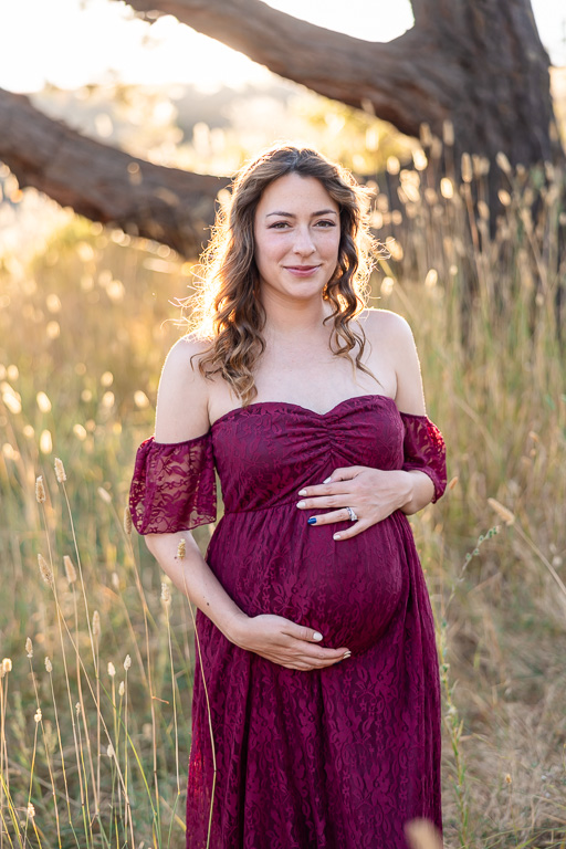 A close-up maternity portrait in Los Altos Hills' Foothills Park at sunset. The pregnant woman is gently holding her baby bump and smiling softly, wearing a burgundy off-shoulder dress in a field of tall grass.