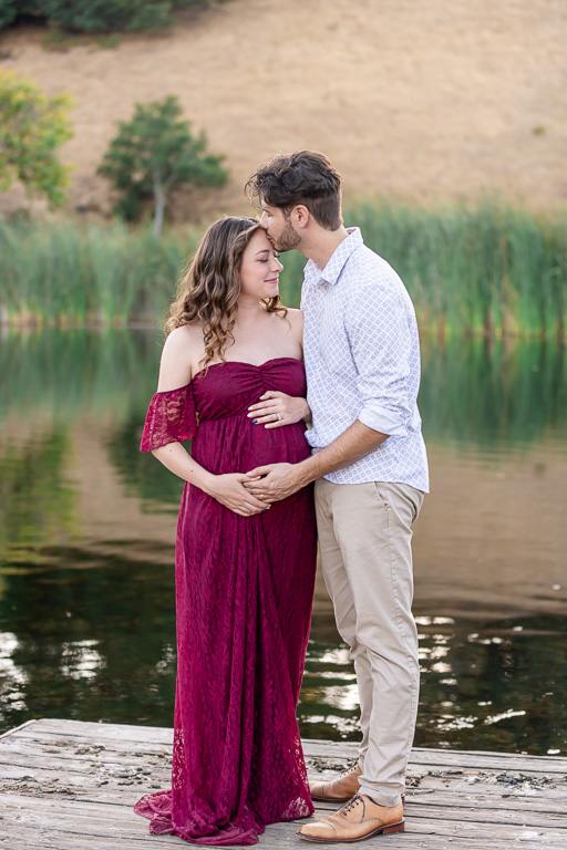 Maternity couple photo on a dock by a lake at Foothills Nature Preserve, Los Altos Hills. The pregnant woman in a burgundy dress and her partner share a quiet moment, their foreheads touching as they stand by the water.