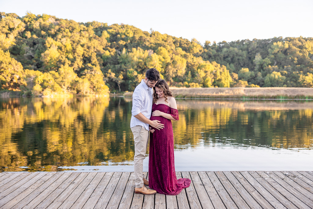 A maternity photo of a couple standing on a dock at Foothills Nature Preserve in Los Altos Hills during sunset. The man is gently holding his partner’s belly as they stand beside a reflective lake surrounded by nature.