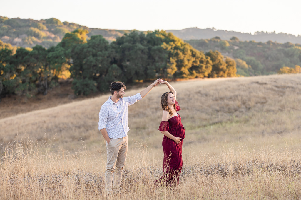 Maternity photo of a couple twirling in an open field at Foothills Park, Los Altos Hills. The pregnant woman’s burgundy maternity dress sways as her partner spins her in the golden light of sunset.