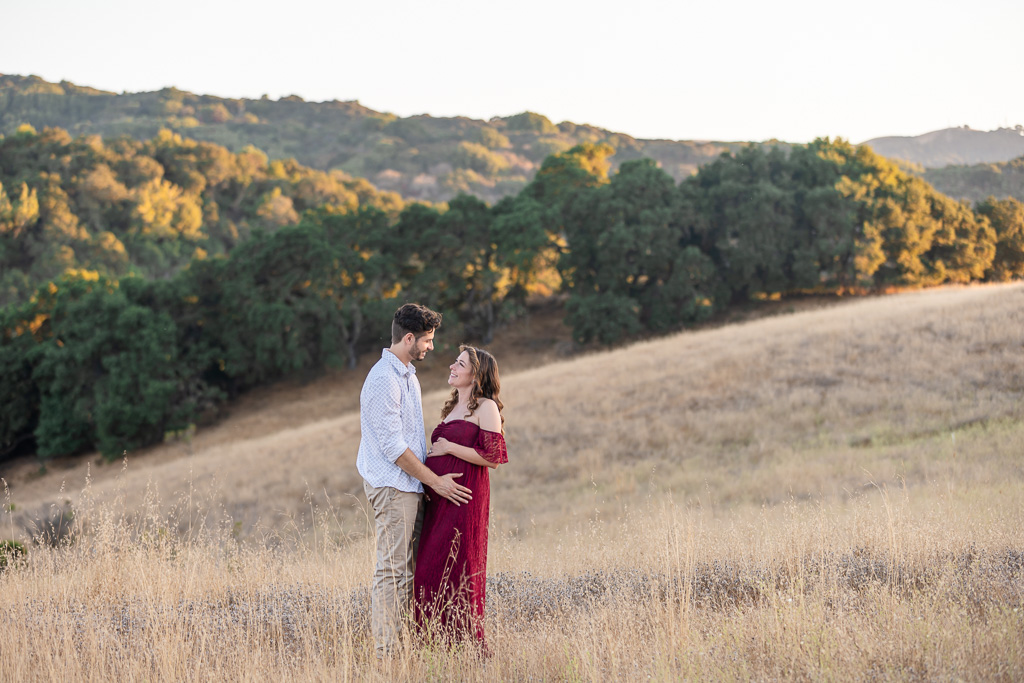 A maternity photo taken in a golden field at Foothills Nature Preserve, Los Altos Hills. The couple is standing face-to-face, smiling at each other, with the woman wearing a burgundy maternity dress and holding her baby bump.