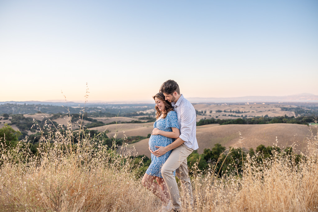 Maternity photo in Foothills Park, Los Altos Hills, overlooking rolling hills at sunset. The pregnant woman in a light blue floral dress stands with her partner, who embraces her from behind as they look at her belly.