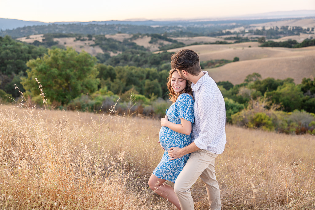 Sunset maternity photo in a dry grassy field at Foothills Nature Preserve, Los Altos Hills. The man kisses the pregnant woman’s forehead as she smiles and holds her baby bump, wearing a light blue floral dress.