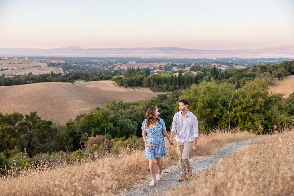 Maternity couple walking hand in hand along a dirt path in Foothills Nature Preserve, Los Altos Hills, with panoramic views of the surrounding hills. The expecting mother is in a light blue maternity dress, and both are smiling at each other.