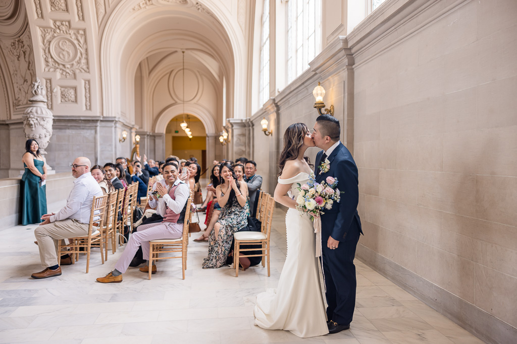 cool shot of bride and groom kissing after wedding ceremony, with guests cheering in the background