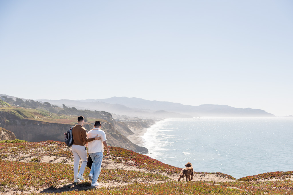 soon-to-be-engaged couple walking with their dog towards a cliff overlooking the ocean and beach below