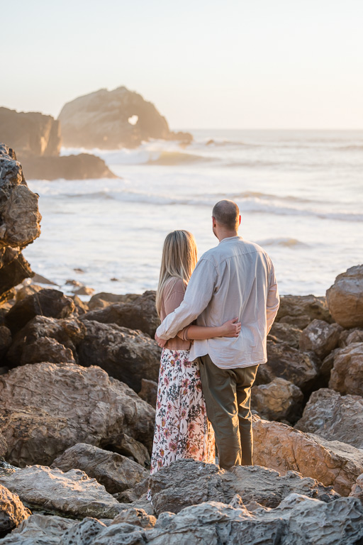 couple standing on rocks by the ocean looking at a heart-shaped hole in the distance