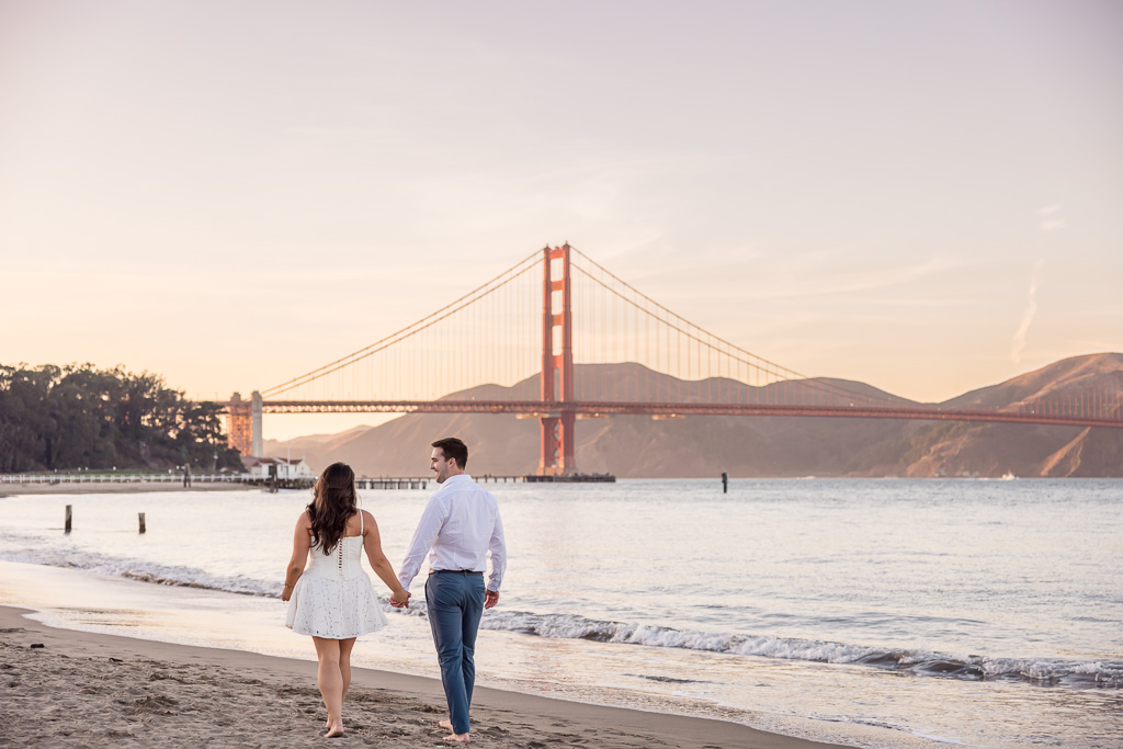 couple on a beach walking towards the Golden Gate Bridge during sunset