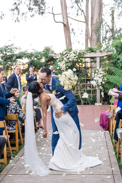 dip & kiss in the middle of the aisle with white flower petals being tossed by guests