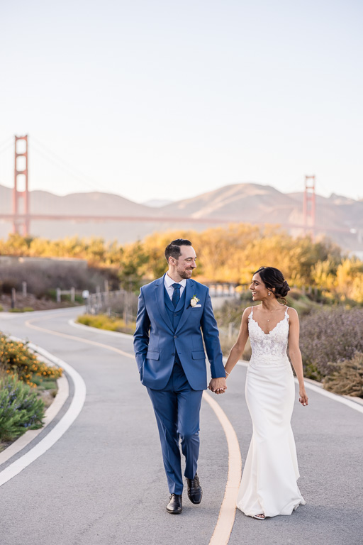 bride and groom walking along a curved road in front of the Golden Gate Bridge at sunset