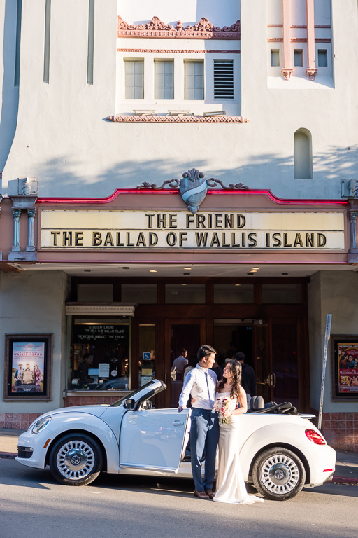 newlywed couple posing with a convertible Beetle under a movie theater marquee