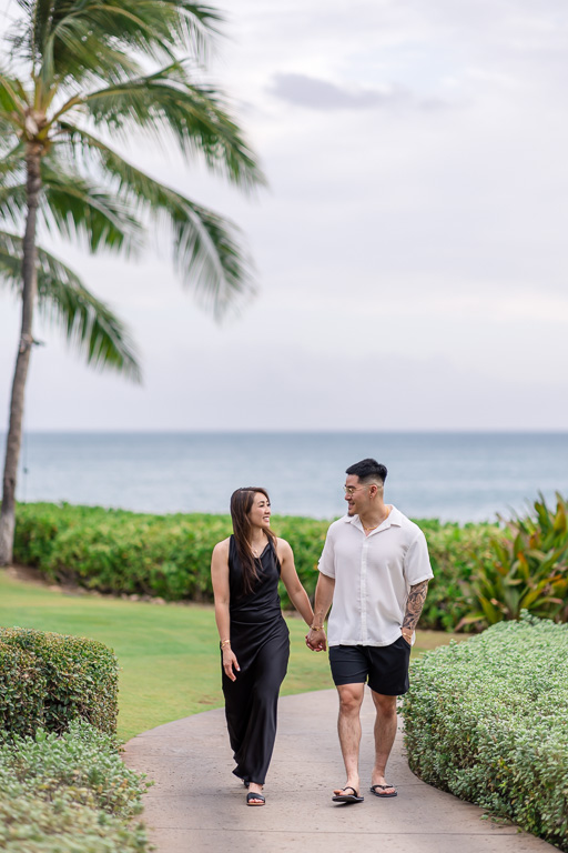 Engaged couple holding hands on a lush green path under towering palm trees in Oahu's Ko Olina resort.