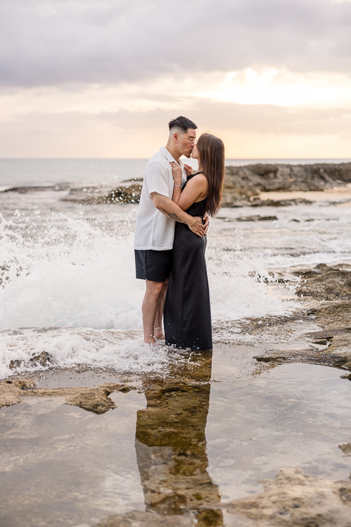 Loving couple embrace in shallow tidal pools at Ko Olina lagoon on Oahu, surrounded by rocky shore and tranquil sunset hues.