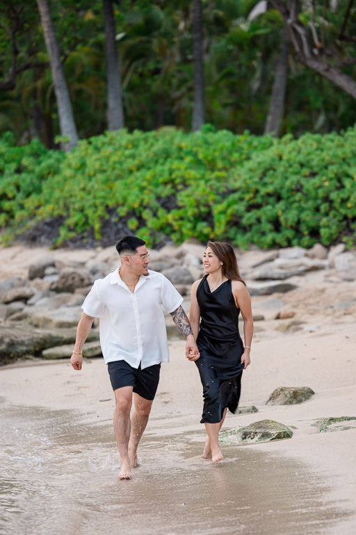 Engaged couple walks hand in hand along the sandy shore of Ko Olina lagoon on Oahu, with lush greenery and palm trees in the background.