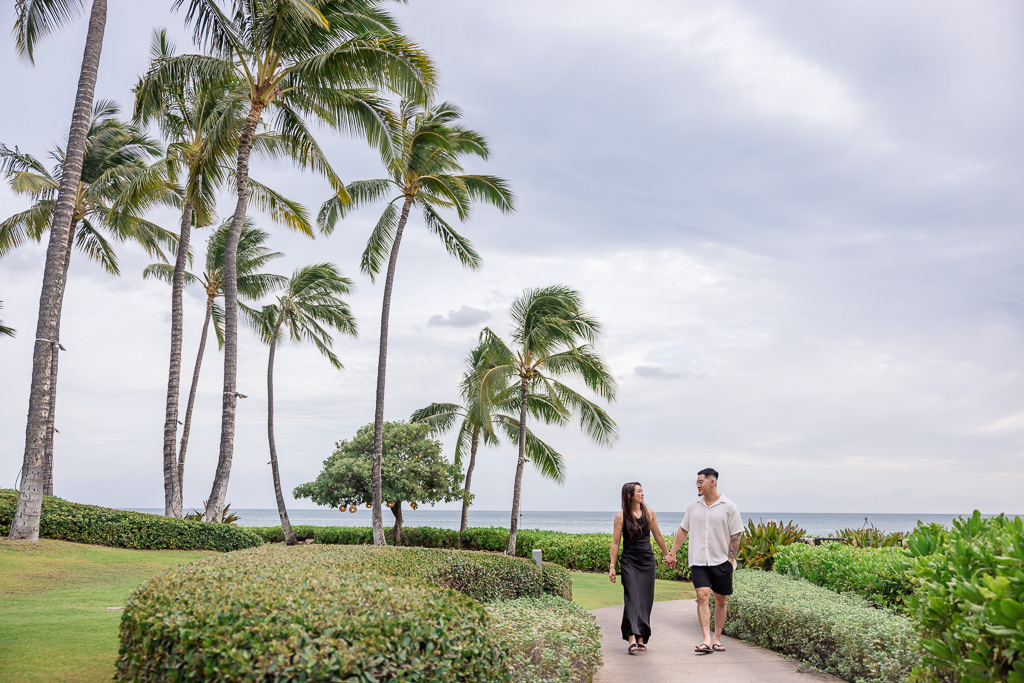 Couple strolls along a paved path lined with tall palm trees in Ko Olina, Oahu, enjoying a tropical lagoon setting.
