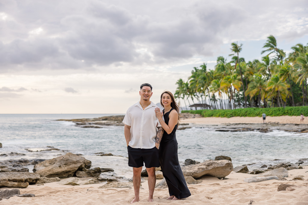 Couple stands together on the sandy shore of Ko Olina lagoon on Oahu with palm trees and blue sky behind them.