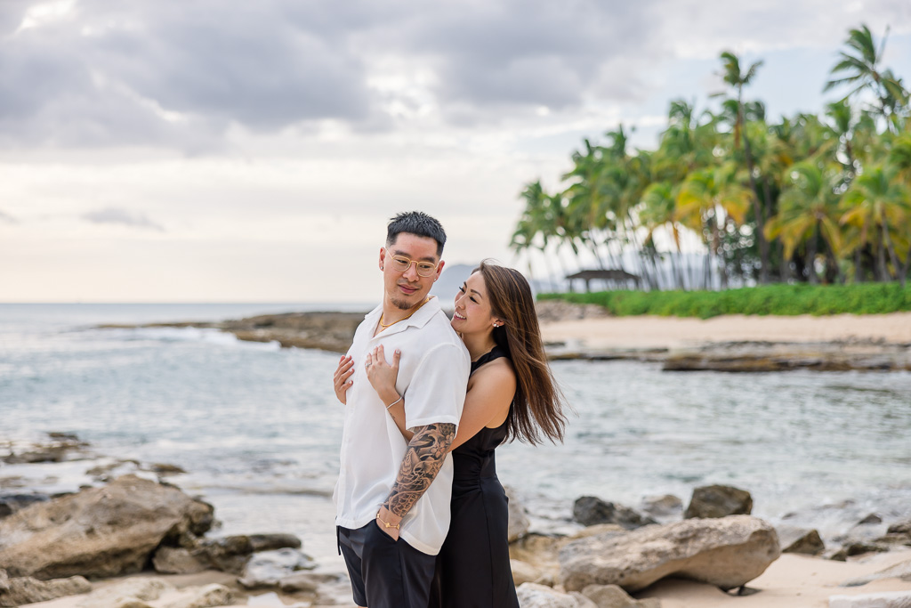 Happy couple hugging by the water's edge at Ko Olina lagoon on Oahu with palm trees and coastal rocks in the background.