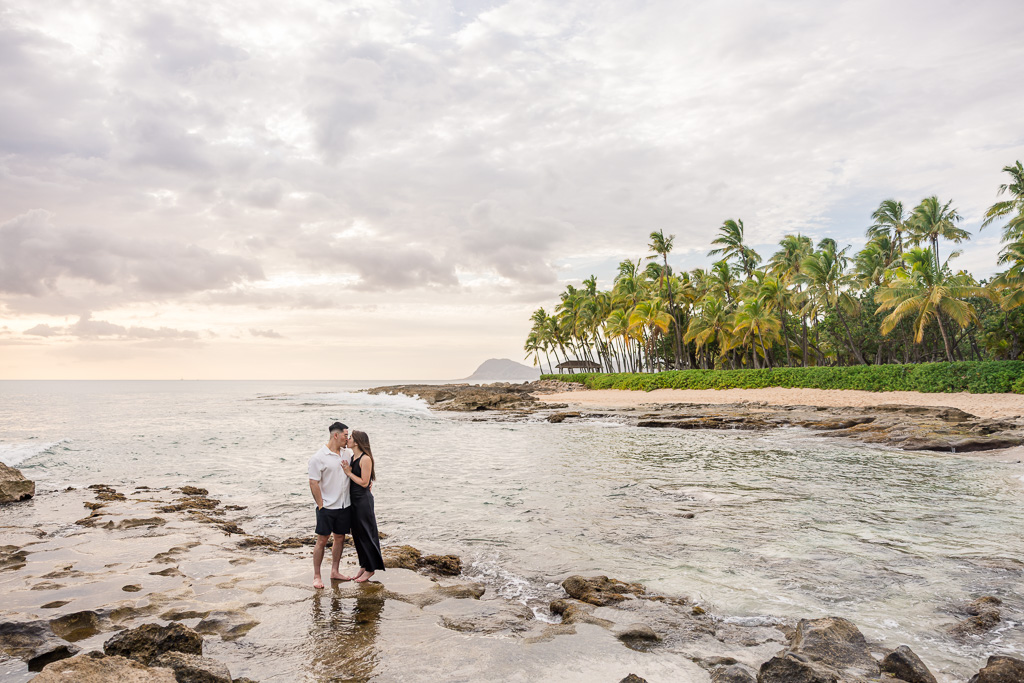 Palm-lined shore of Ko Olina lagoon on Oahu at sunset with golden light reflecting in the water and a couple standing on the rocks.