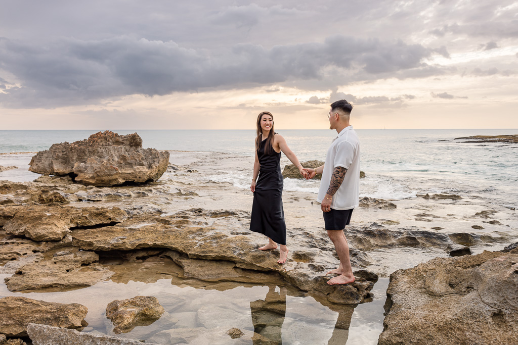 Couple strolls hand in hand along the rocky shoreline of Ko Olina lagoon on Oahu as the sun sets over the ocean.
