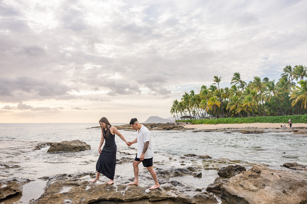 Playful couple shares a moment on the rocky edge of Ko Olina lagoon with a backdrop of calm ocean waters, palm trees and pastel sunset on Oahu.