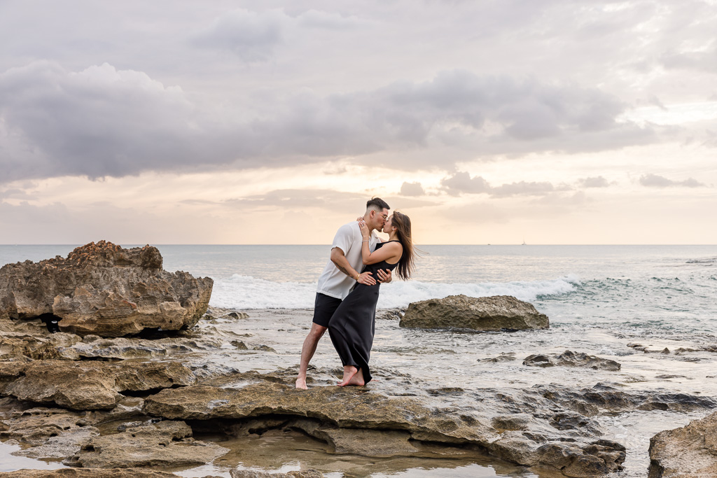 Engaged couple poses together on the rocky shoreline of Ko Olina lagoon in Oahu, with ocean waves and a pastel sunset sky behind them.